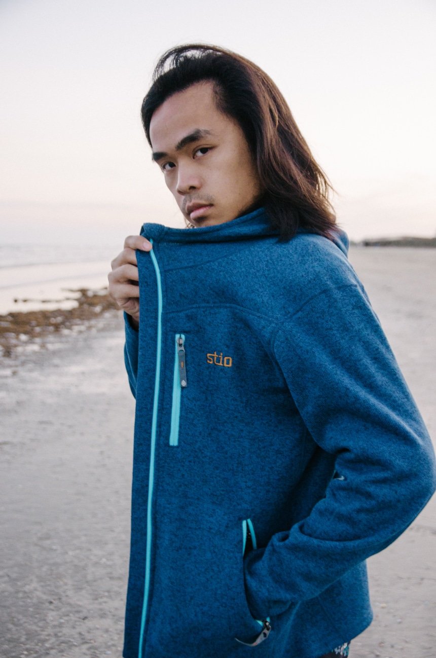 Close Up On Logo Of Model Wearing Stio Wilcox Fleece Hoodie and Looking In Camera At The Beach