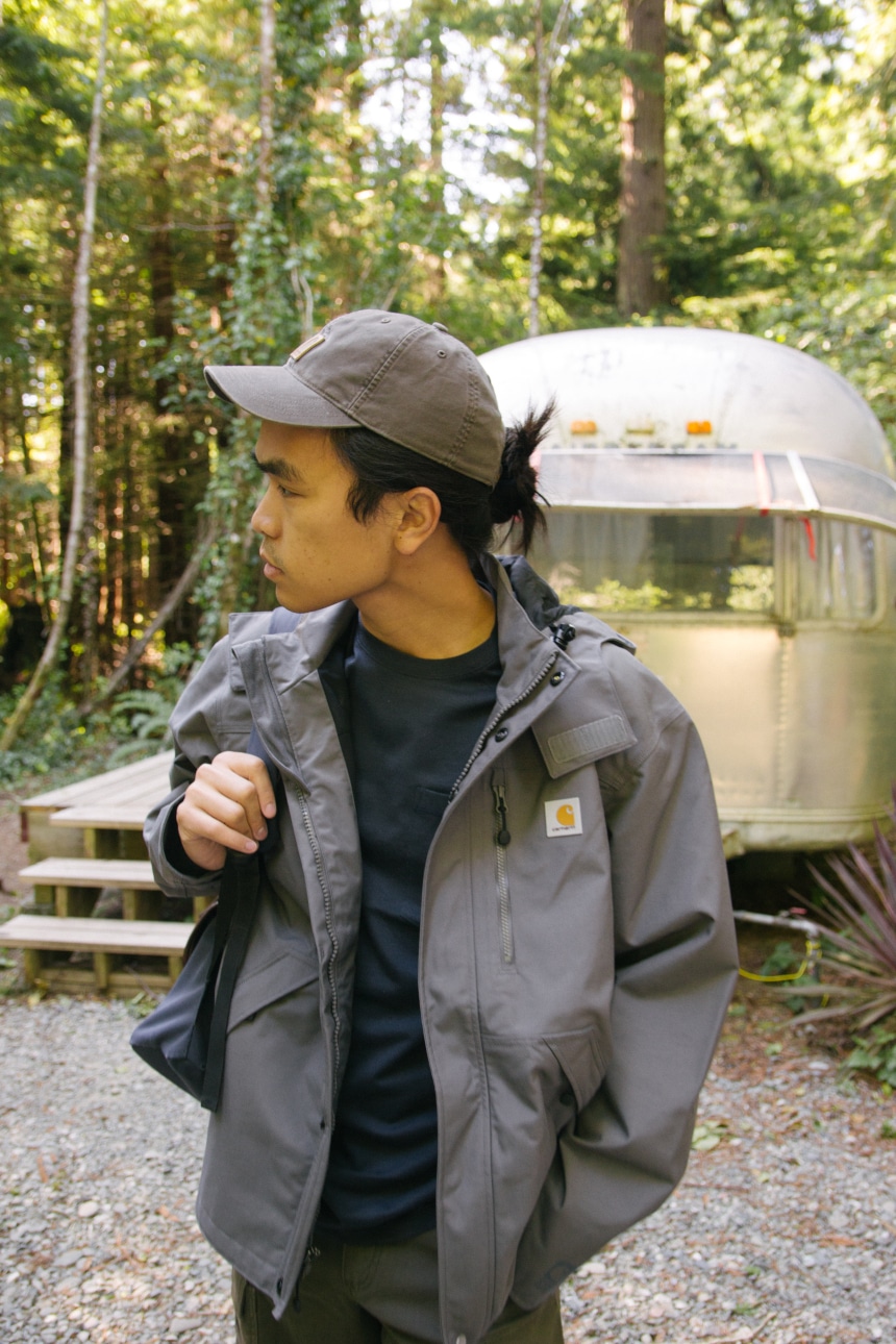 Man in woods holding backpack looking down wearing Carhartt Odessa Cap Rugged Flex Rigby Dungaree and Force Delmont Tee Looking to the Right