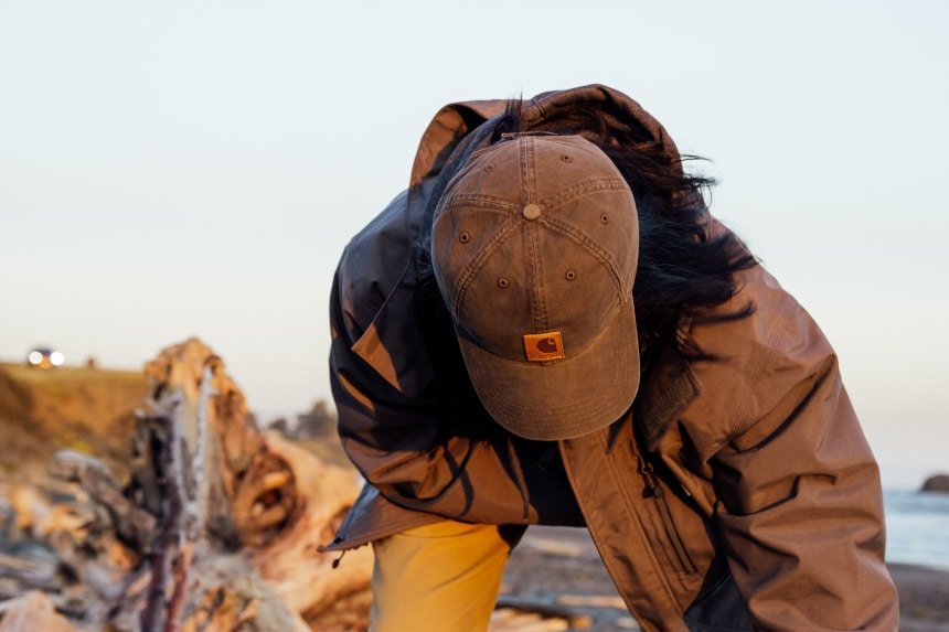 Man on beach picking up driftwood wearing Carhartt Shoreline Jacket Odessa Cap and Rugged Flex Rigby Dungaree