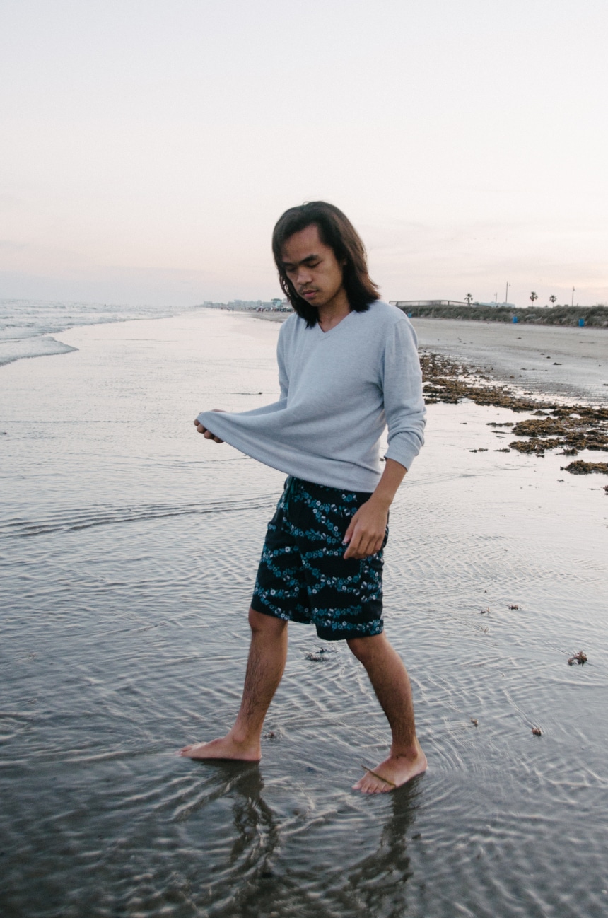 Model Wearing Stio CFS Board Short While Walking In The Water At The Beach And Holding Up Shirt