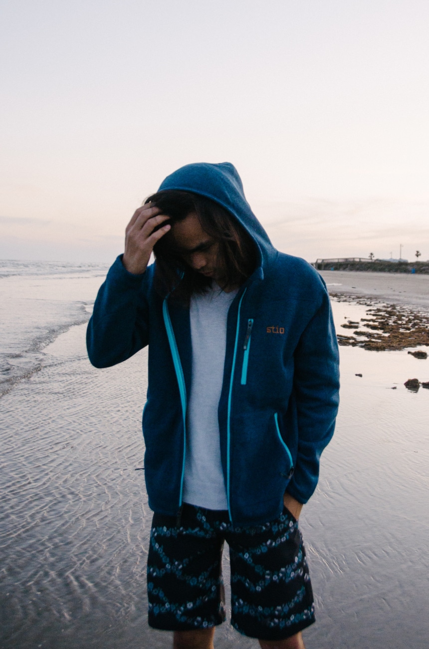 Model Wearing Stio Wilcox Fleece Hoodie and CFS Board Shorts While Holding Hood at the Beach While Looking Down