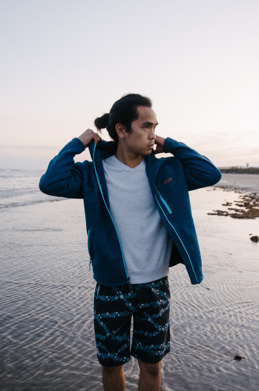 Model Wearing Stio Wilcox Fleece Hoodie and CFS Board Shorts While Holding Hood at the Beach While Looking In The Distance